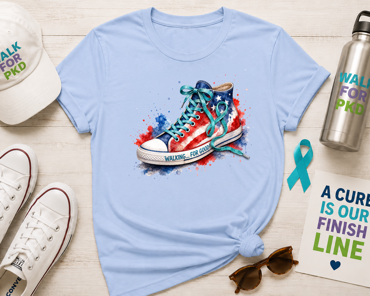 A flat-lay of a light blue t-shirt featuring a patriotic sneaker graphic with 'WALKING FOR GOOD' text, styled with a white 'WALK FOR PKD' cap, white Converse sneakers, a silver water bottle, sunglasses, and a support card on a light wood background.